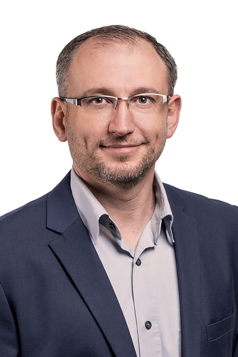 Andrej Justus, verantwortlich für den Vertrieb von Systemböden und das Produktionsmanagement, mit Brille und leichtem Bart – lächelt leicht, trägt einen blauen Blazer und ein hellgraues Hemd vor weißem Hintergrund. Professioneller und freundlicher Ton.