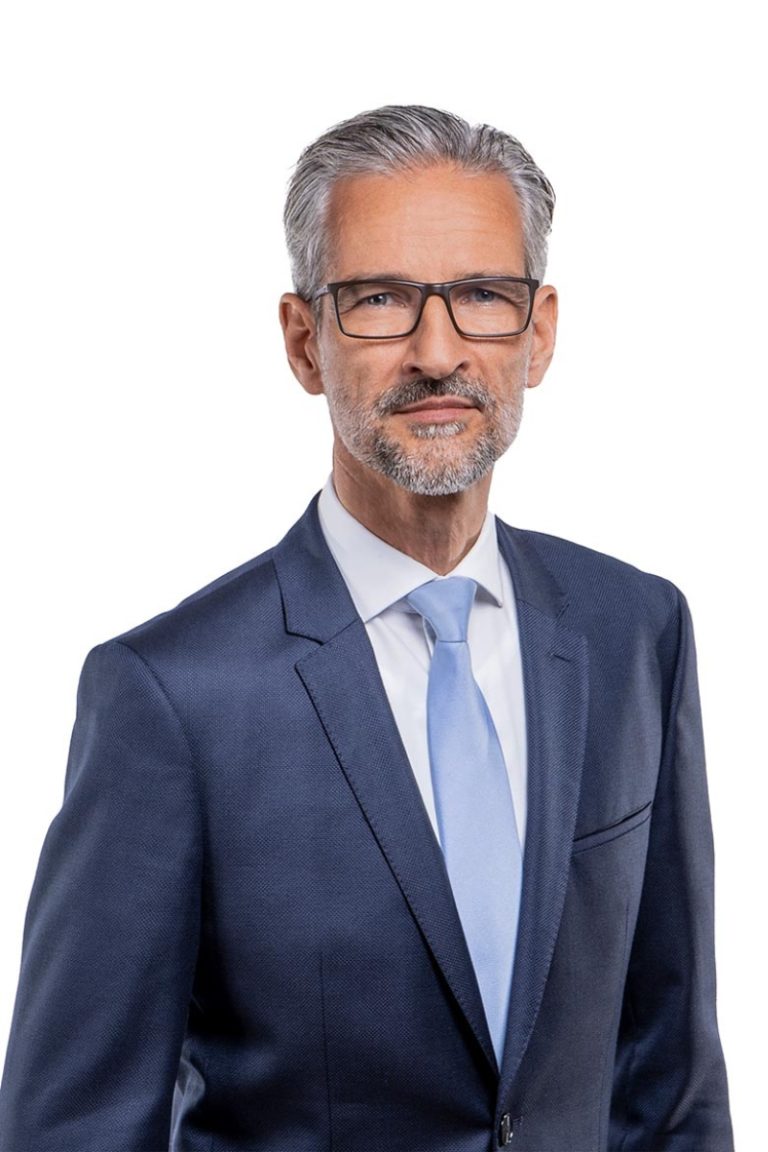 Geschäftsführer Marc Friedrich, mit grauen Haaren und Bart, Brille, dunkelblauem Anzug und hellblauer Krawatte, steht vor einem weißen Hintergrund und strahlt Professionalität aus.
